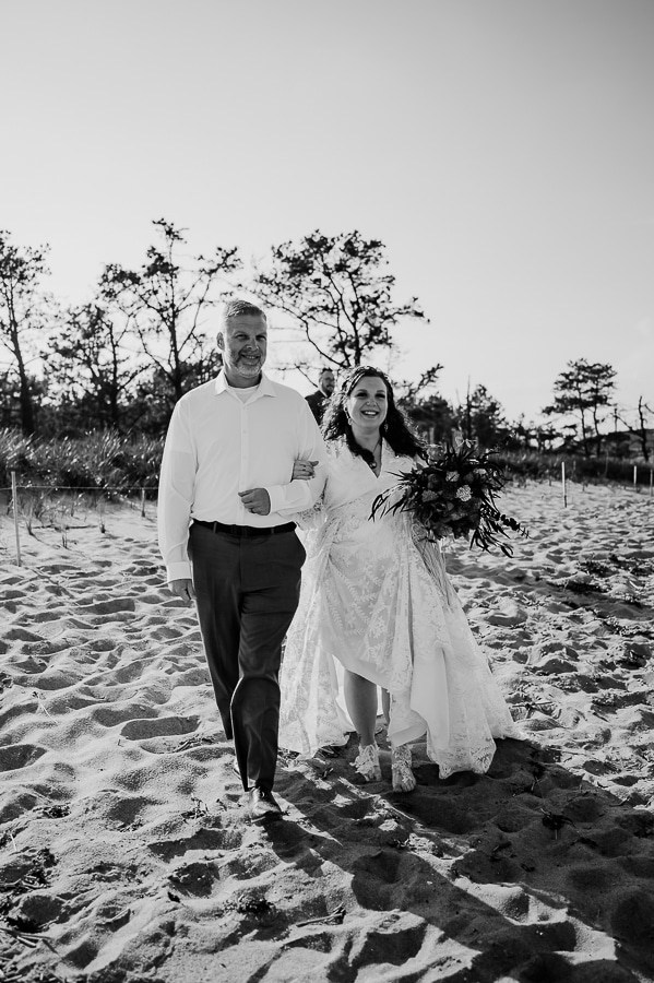 Black and white father walking bride across beach to wedding ceremony