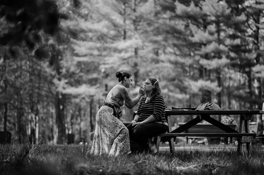 Black and white bride getting makeup done while sitting on picnic table in woods