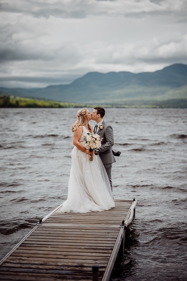 Bride and groom kissing on end of dock on lake with mountains