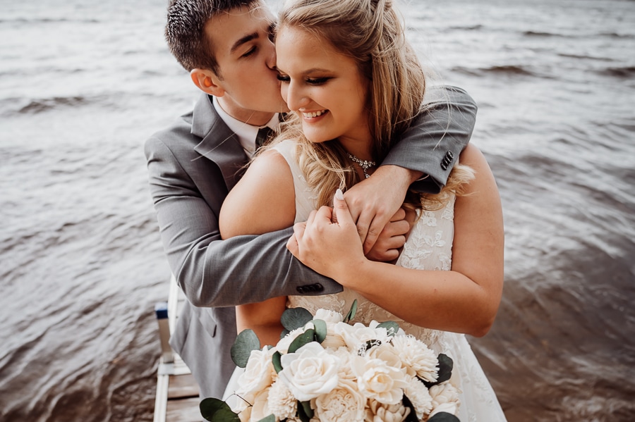 Groom wrapping arms over brides shoulders in front of the lake