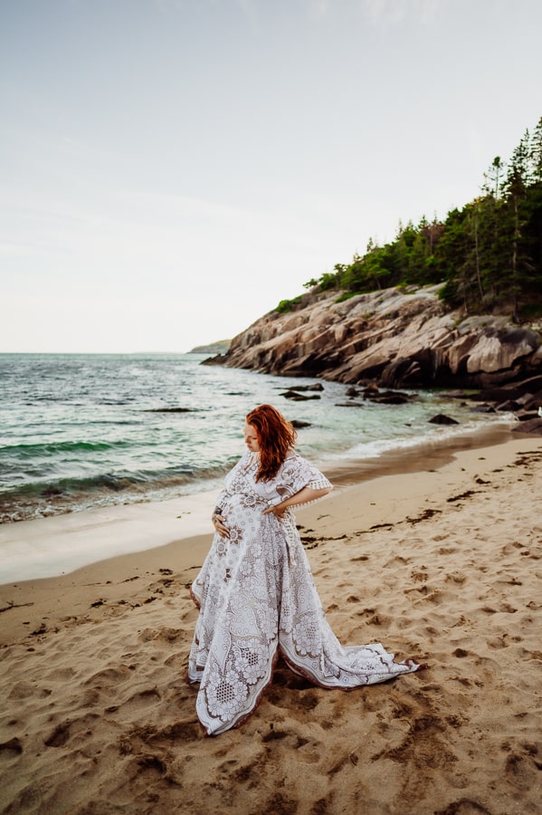 Woman posing in front of ocean in white lace dress for maternity photos