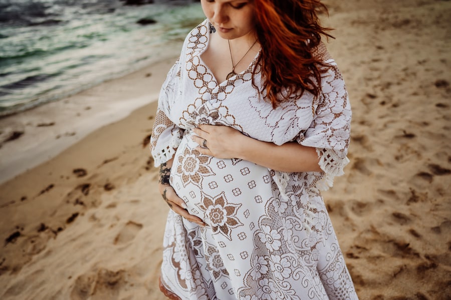 Woman in front of ocean on sand beach in white maternity dress