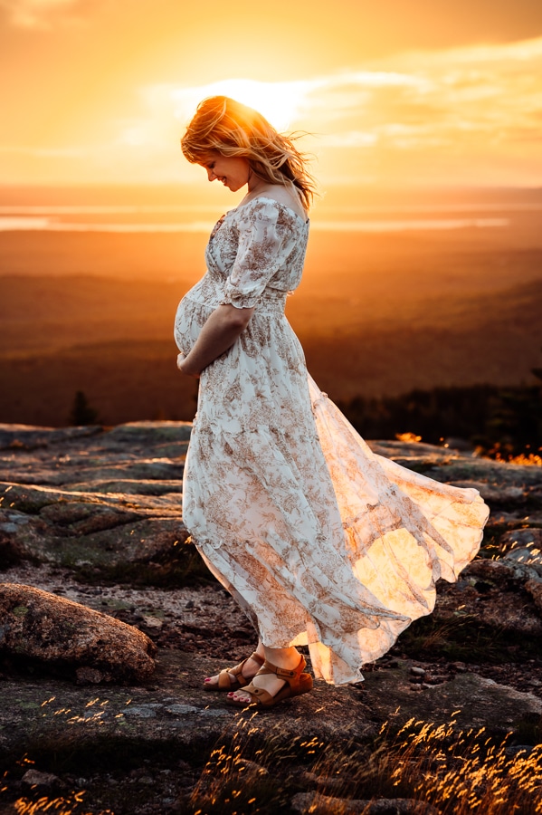 Woman in white dress with brown florals holding belly on cadillac mountain at sunset