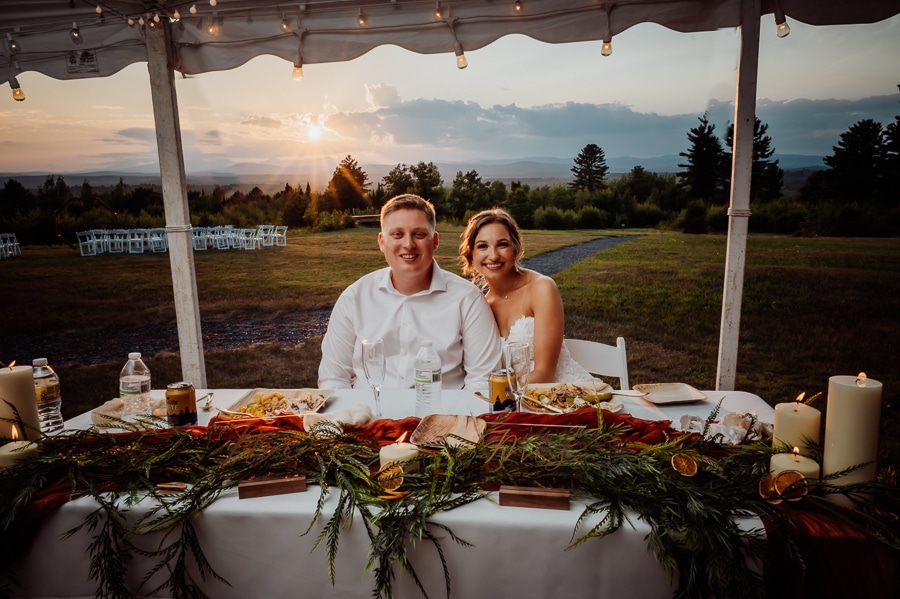 Bride and groom sitting at bridal table under tent with sunset and chairs in background
