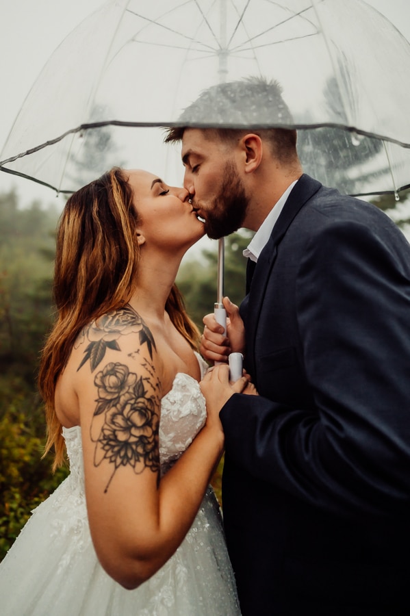 Bride with arm tattoo with groom under clear umbrella kissing in rain