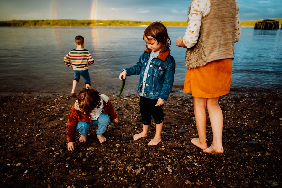 Kids in bold colors picking up shells and seaweed on beach in front of double rainbow