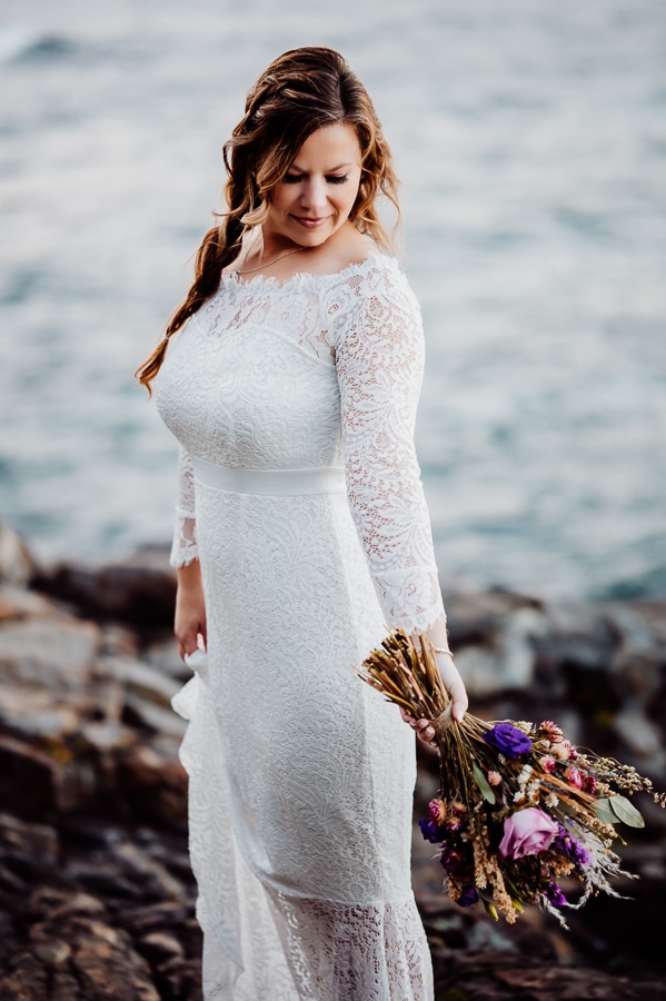 Bride looking down at shoulder with flowers upside down in front of ocean in Maine