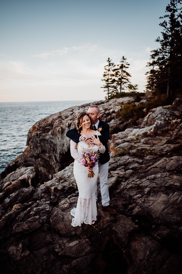 Groom with arms wrapped around brides shoulders kissing her cheek on Ocean cliffs