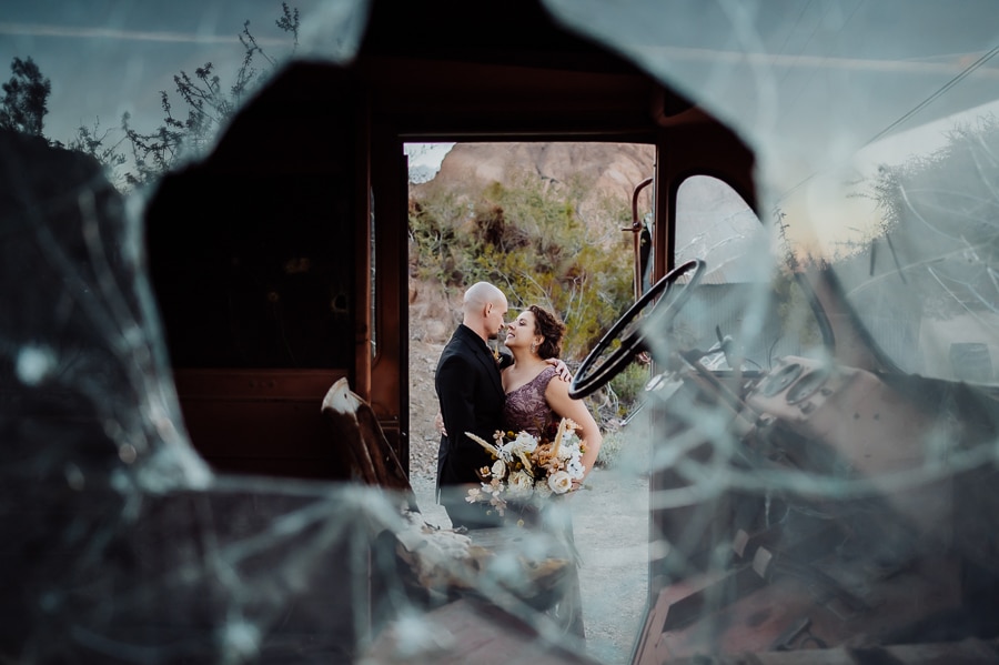 bride and groom kissing by old truck framed with broke glass window